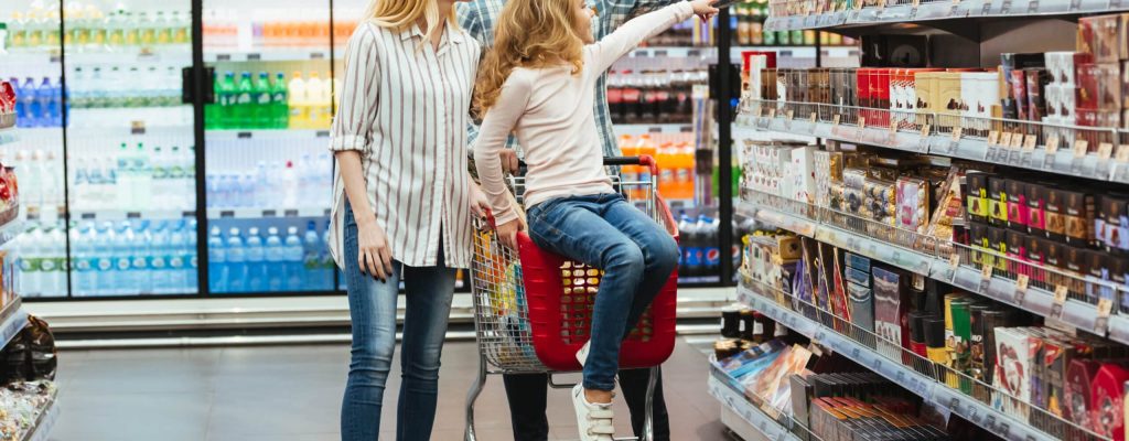 Cheery little girl sitting on a shopping cart and choosing candy with her parents at the supermarket